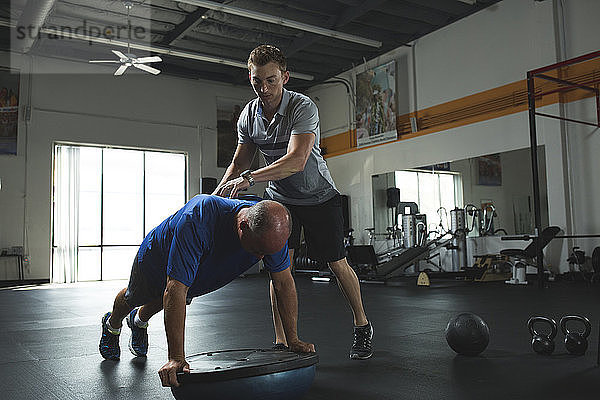 Trainer hilft dem Kunden beim Üben von Liegestützen auf dem Bosu-Ball mit Gleichgewichtsscheibe im Fitnessstudio