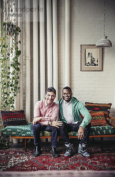 Ganzfigur eines glücklichen multiethnischen schwulen Paares  das zu Hause auf einer Chaiselongue sitzt