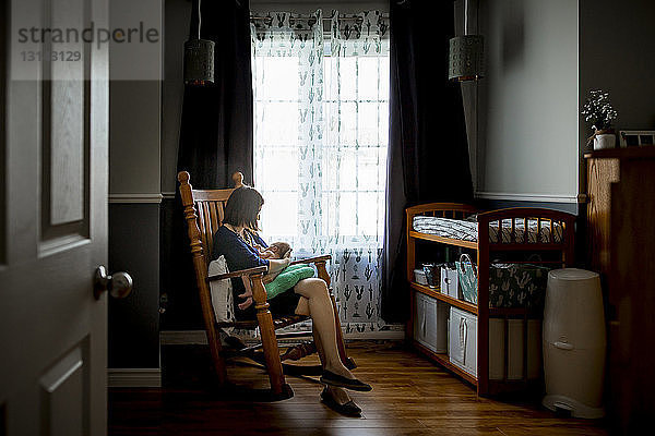 Mutter stillt neugeborenen Sohn  während sie auf einem Schaukelstuhl sitzt und durch die Tür zu Hause gesehen wird