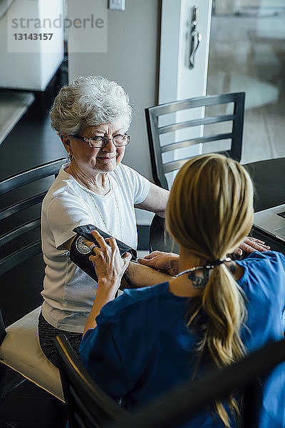 Schrägansicht der häuslichen Pflegekraft beim Einstellen des Blutdrucks Druckmesser am Arm einer älteren Frau am Esstisch