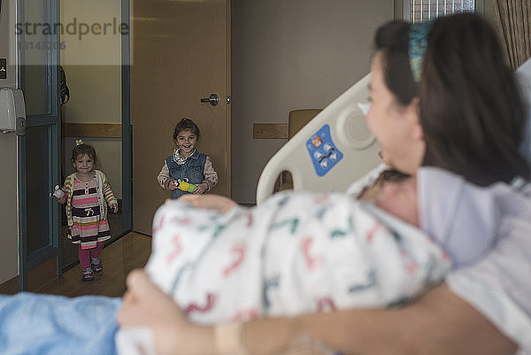 Mutter hält neugeborenen Sohn und sieht Töchter  die in die Krankenhausstation kommen  während sie sich auf dem Bett ausruht