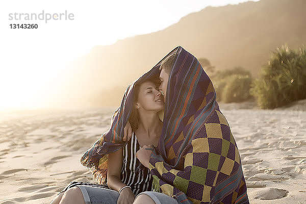 Junges Paar küsst sich am Strand bei Sonnenuntergang  während es in eine Decke gehüllt ist
