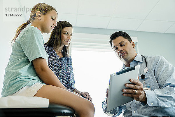 Niedrigwinkelansicht eines Kinderarztes  der Mutter und Tochter im medizinischen Untersuchungsraum einen Tablet-Computer zeigt