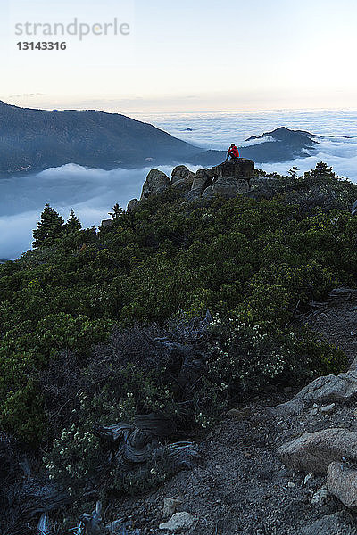 Fernansicht einer Frau  die auf dem Gipfel eines Berges in einer Wolkenlandschaft sitzt