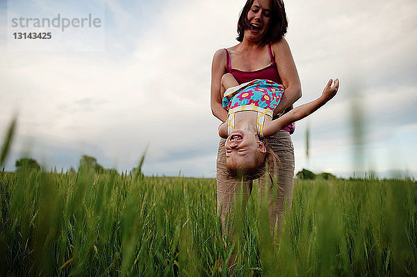 Tiefwinkelansicht einer verspielten Mutter  die ihre Tochter kopfüber auf einem Grasfeld vor bewölktem Himmel trägt