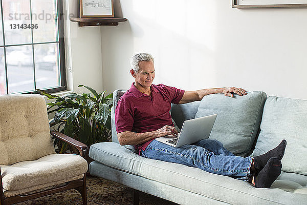 Reifer Mann in voller Länge mit Laptop-Computer  während er zu Hause auf dem Sofa im Wohnzimmer ruht