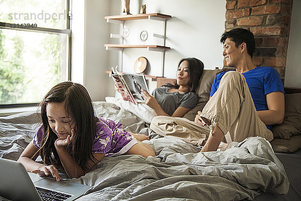 Mädchen benutzt Laptop-Computer  während sie sich zu Hause mit den Eltern im Bett entspannt