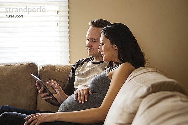 Mann benutzt Tablet-Computer  während schwangere Frau zu Hause auf dem Sofa sitzt