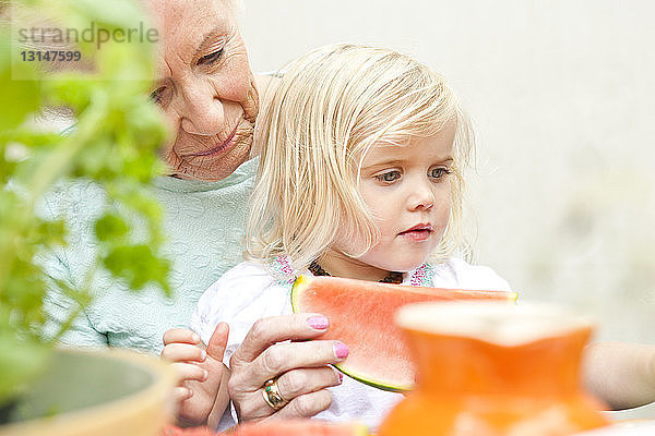 Ältere Frau und kleine Urenkelin essen Melonenstückchen am Gartentisch