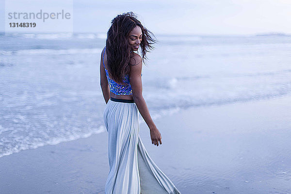 Junge Frau geht am Strand spazieren  schaut über die Schulter  lächelt