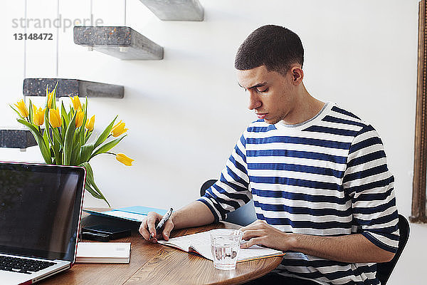 Junger Mann sitzt am Tisch und schreibt in ein Notizbuch