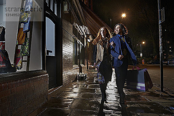 Vorderansicht eines Paares  das Arm in Arm eine Straße entlangläuft und eine Einkaufstasche bei Nacht trägt  Schaufensterbummel