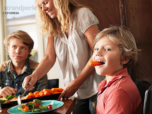 Junge hält Karotte im Mund  während Mutter gesundes Essen serviert
