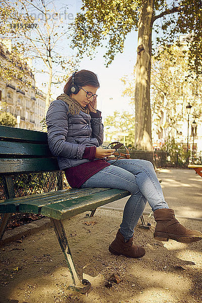 Mittlere erwachsene Frau sitzt im Park und wählt auf ihrem Smartphone Musik für Kopfhörer aus