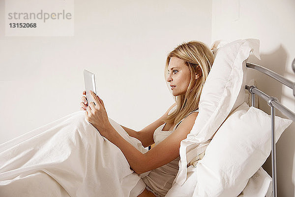 Mittlere erwachsene Frau  die im Bett sitzt und ein digitales Tablet benutzt