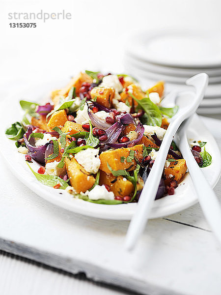 Schneidebrett mit Butternusskürbis-Salat  Feta  roten Zwiebeln  Granatapfel und grünen Salatblättern auf dem Gartentisch