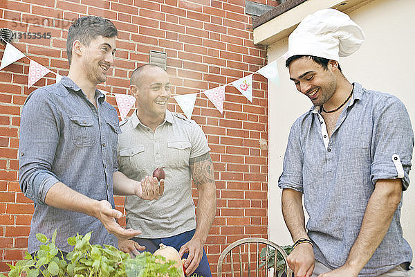 Männliche Freunde lachen über die Kochmütze  während sie Essen für ein Grillfest im Garten vorbereiten