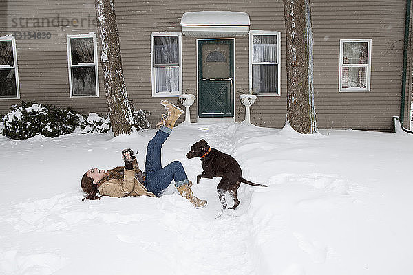 Mittlere erwachsene Frau liegt im Schnee vor dem Haus und spielt mit einem Hund