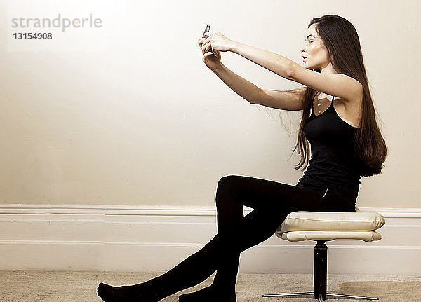 Seitenansicht einer jungen Frau  die auf einem Hocker sitzt und ein Smartphone benutzt  um ein Selfie zu machen