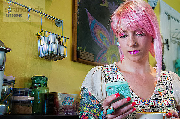 Junge Frau mit rosa Haaren liest in der Küche eine Textnachricht auf dem Smartphone