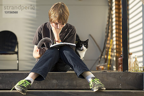 Junge sitzt auf einer Stufe und liest ein Buch  die Katze schaut von hinten zu