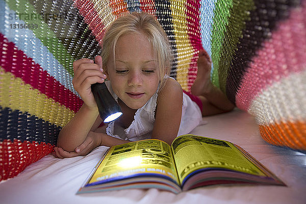 Auf dem Bett liegendes Mädchen  das sich unter einer Decke versteckt und mit einer Taschenlampe eine Zeitschrift liest