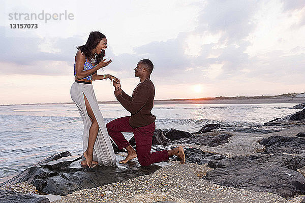 Mittlerer erwachsener Mann kniet auf Felsen am Meer und macht einer jungen Frau einen Heiratsantrag