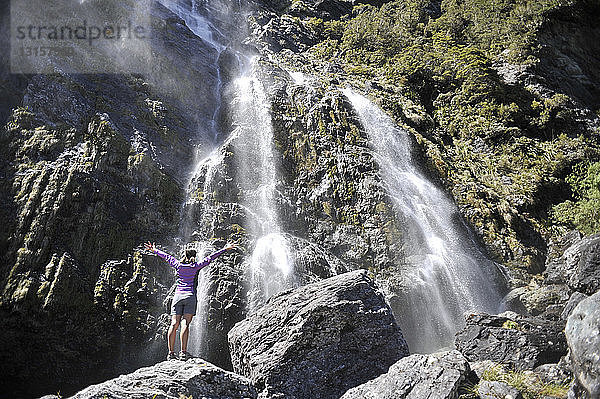 Frau steht mit ausgestreckten Armen am Wasserfall  Neuseeland