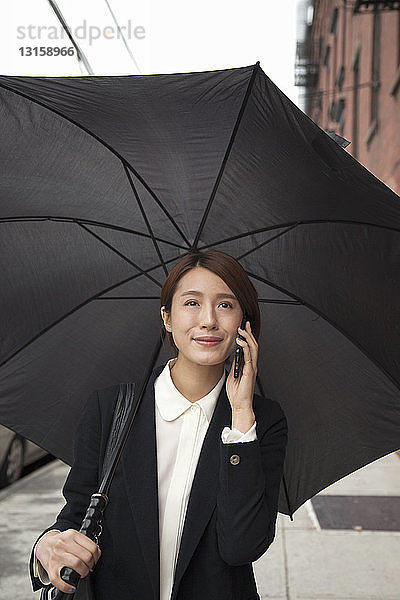 Geschäftsfrau mit Regenschirm  die auf der Straße ihr Smartphone benutzt