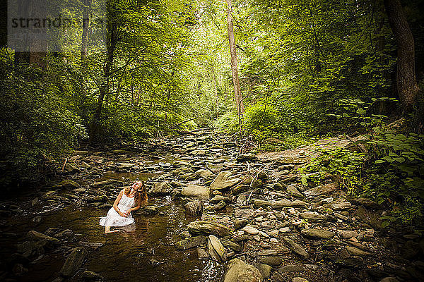 Mittlere erwachsene Frau in weißem Kleid sitzt auf einem Felsen im Flussbett und schaut nach oben