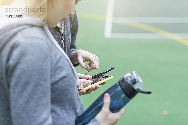 Abgeschnittene Seitenansicht einer jungen Frau auf einem Sportplatz  die eine Wasserflasche hält und auf ihr Smartphone blickt