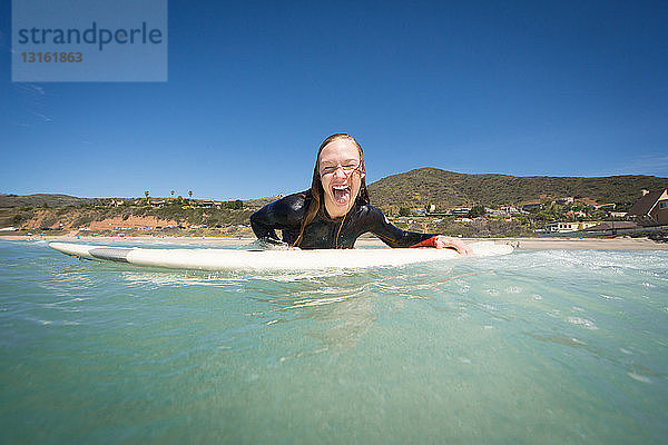 Vorderansicht einer jungen Frau  die auf einem Surfbrett liegt und in die Kamera schaut und ihre Zunge herausstreckt. Los Angeles  Kalifornien  USA