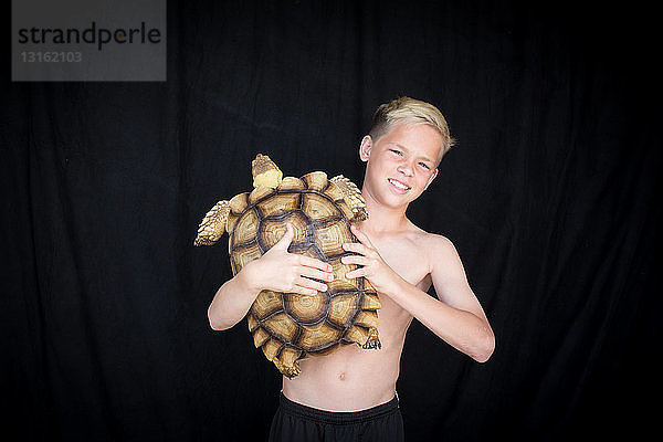 Vorderansicht eines Jungen mit nacktem Oberkörper  der eine afrikanische Spornschildkröte hält  in die Kamera blickend