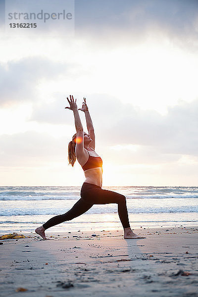 Mittlere erwachsene Frau  die bei Sonnenuntergang am Strand eine Krieger-Yoga-Pose einnimmt