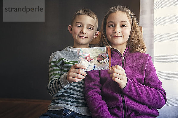 Porträt von Zwillingsjungen und -mädchen  die ein Foto von sich selbst als Babys hochhalten