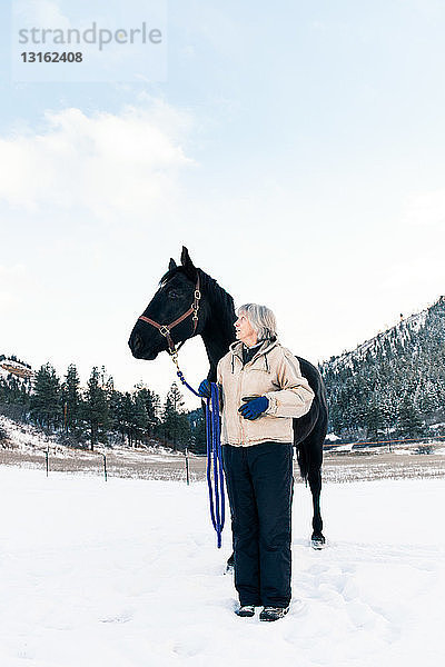 Ältere erwachsene Frau steht mit Pferd in verschneiter Landschaft