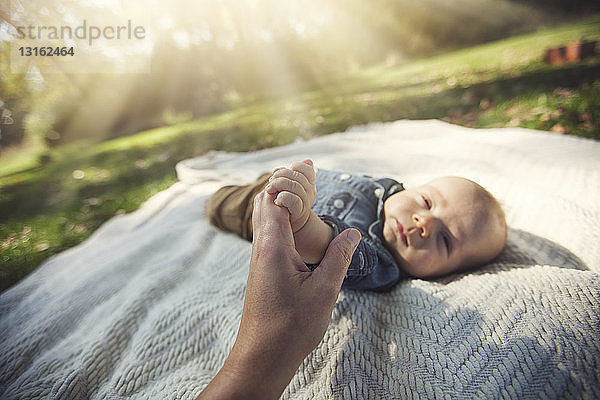 Mutter hält Hand eines kleinen Jungen  der im Sonnenlicht auf einer Decke liegt und in die Kamera schaut