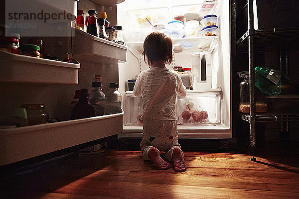 Rückansicht eines männlichen Kleinkindes  das nachts vor einem offenen Kühlschrank kniet