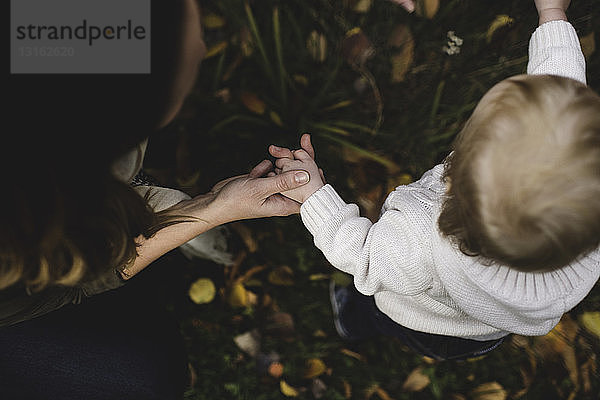 Geschnittene Draufsicht von Mutter und Kind  die auf Herbstblättern stehen und sich an den Händen halten