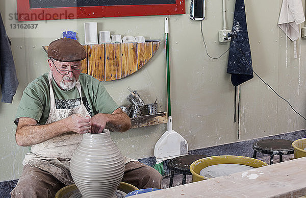 Töpfer mit flacher Kappe sitzt an einer Tonvase  die eine Töpferscheibe formt