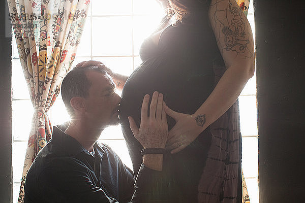 Schrägansicht einer tätowierten schwangeren  mittleren erwachsenen Frau  Partner kniend und den Bauch küssend
