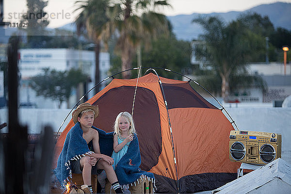 Bruder und Schwester mit Ghettoblaster sitzen neben einem orangefarbenen  in eine Decke gehüllten Zelt