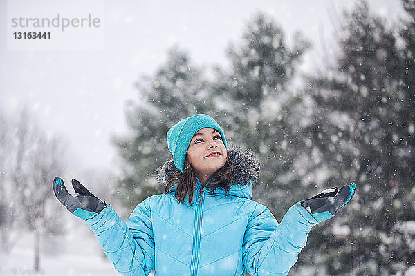Mädchen mit türkisfarbener Strickmütze und -mantel  die Arme erhoben  die Hände nach Schnee streckend  lächelnd aufblickend