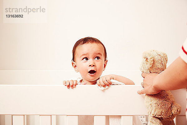 Baby schaut über das Kinderbett  schaut die Mutter mit Plüschtier an