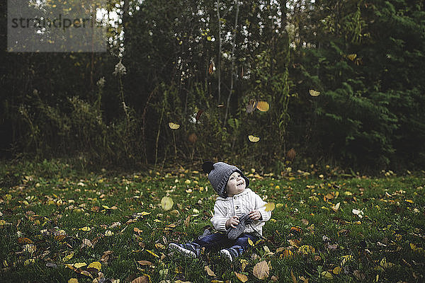 Kleiner Junge mit Pudelmütze sitzt im Gras und beobachtet lächelnd das fallende Herbstlaub