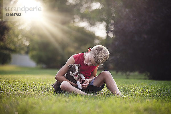 Frontansicht eines Jungen  der mit einem Boston-Terrier-Welpen im Gras sitzt und nach unten schaut