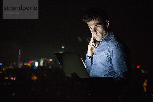 Junger Geschäftsmann schaut nachts vor dem Fenster eines Bürohochhauses auf seinen Laptop  Shanghai  China