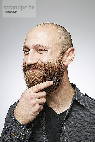 Studioporträt eines mittelgroßen erwachsenen Mannes mit rasiertem Haar  das über den überwachsenen Bart streicht