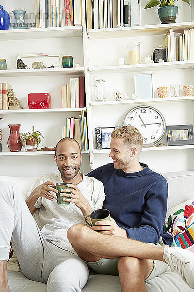 Homosexuelles Paar sitzt auf dem Sofa  hält Kaffeetassen in der Hand und schaut lächelnd in die Kamera