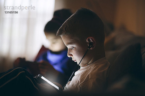 Seitenansicht eines Jungen auf dem Sofa  der Kopfhörer trägt und mit einem digitalen Tablet nach unten schaut